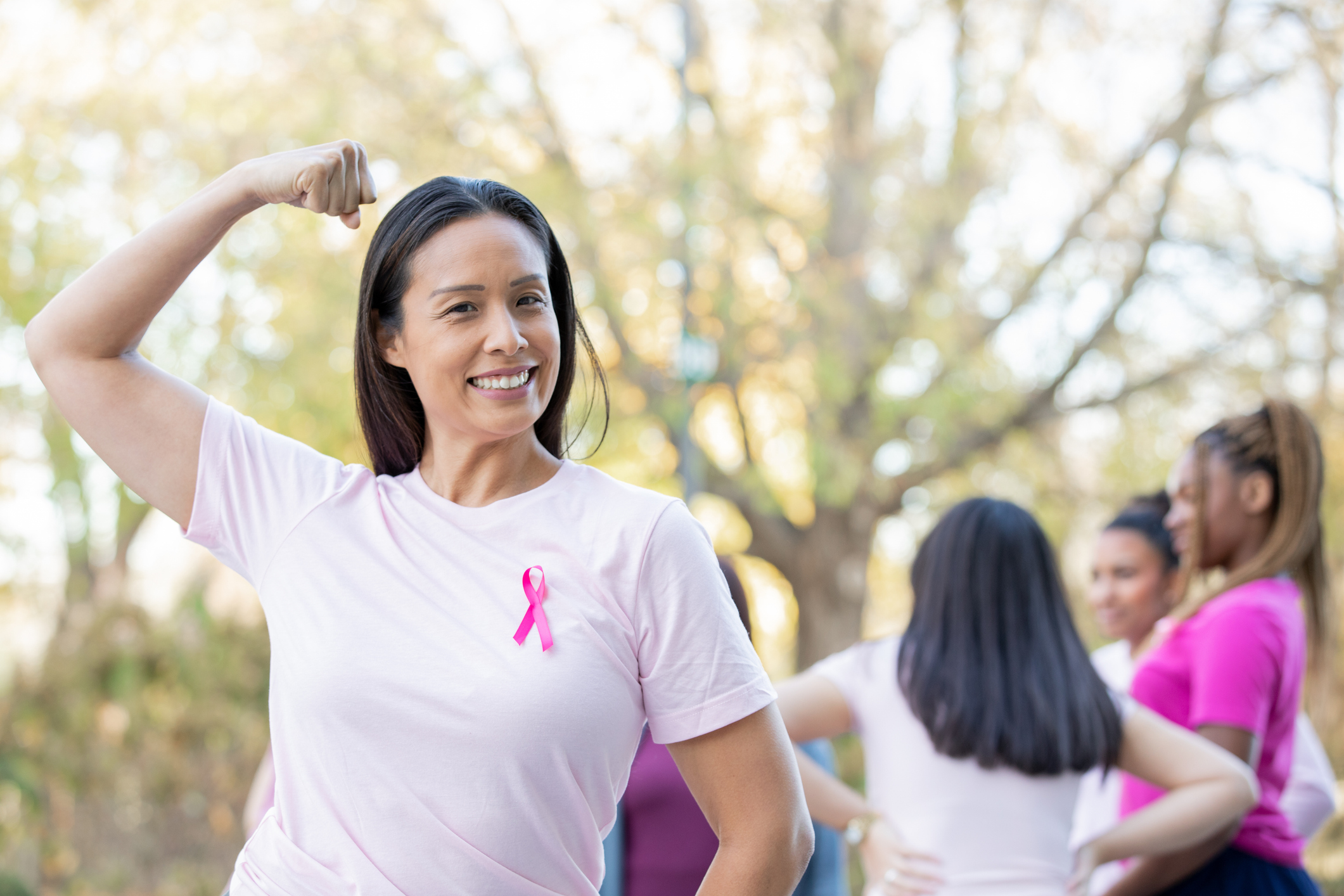 Mulher faz sinal de força, ao mostrar o seu músculo do braço, e veste um camiseta que tem o símbolo da luta contra o câncer de mama