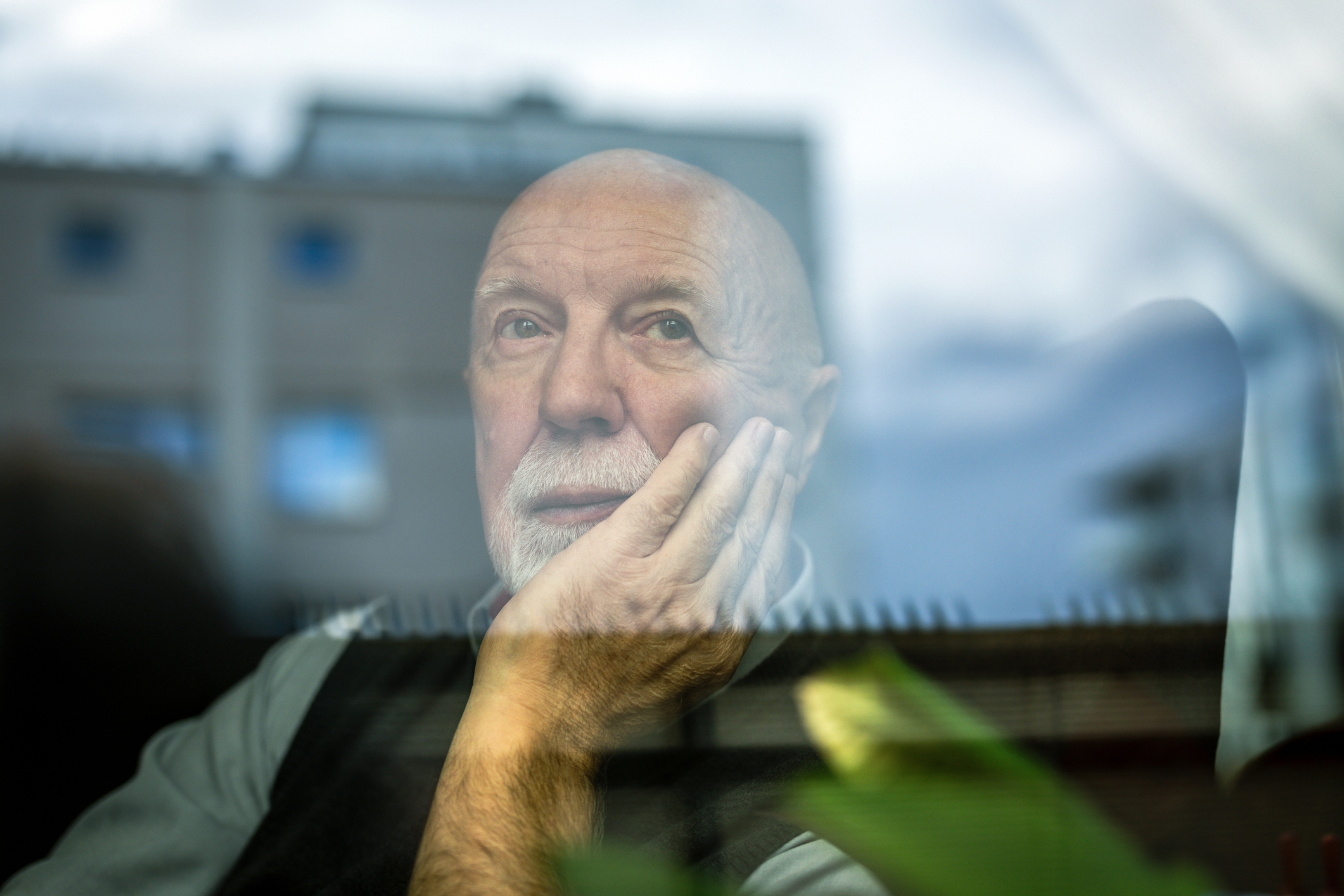 Senhor com mão segurando o queixo olha pela janela sereno. O alzheimer é uma doença que comete muitas pessoas da terceira idade